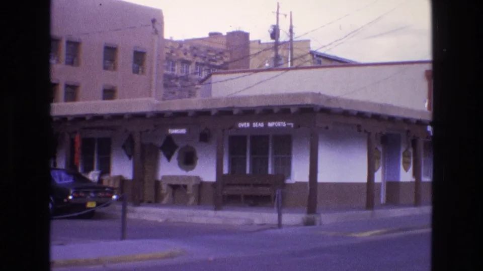 1973 New Mexico street scene with a low commercial building, covered walkway, shop signage, utility lines, and the rear of a dark parked car at the left edge.