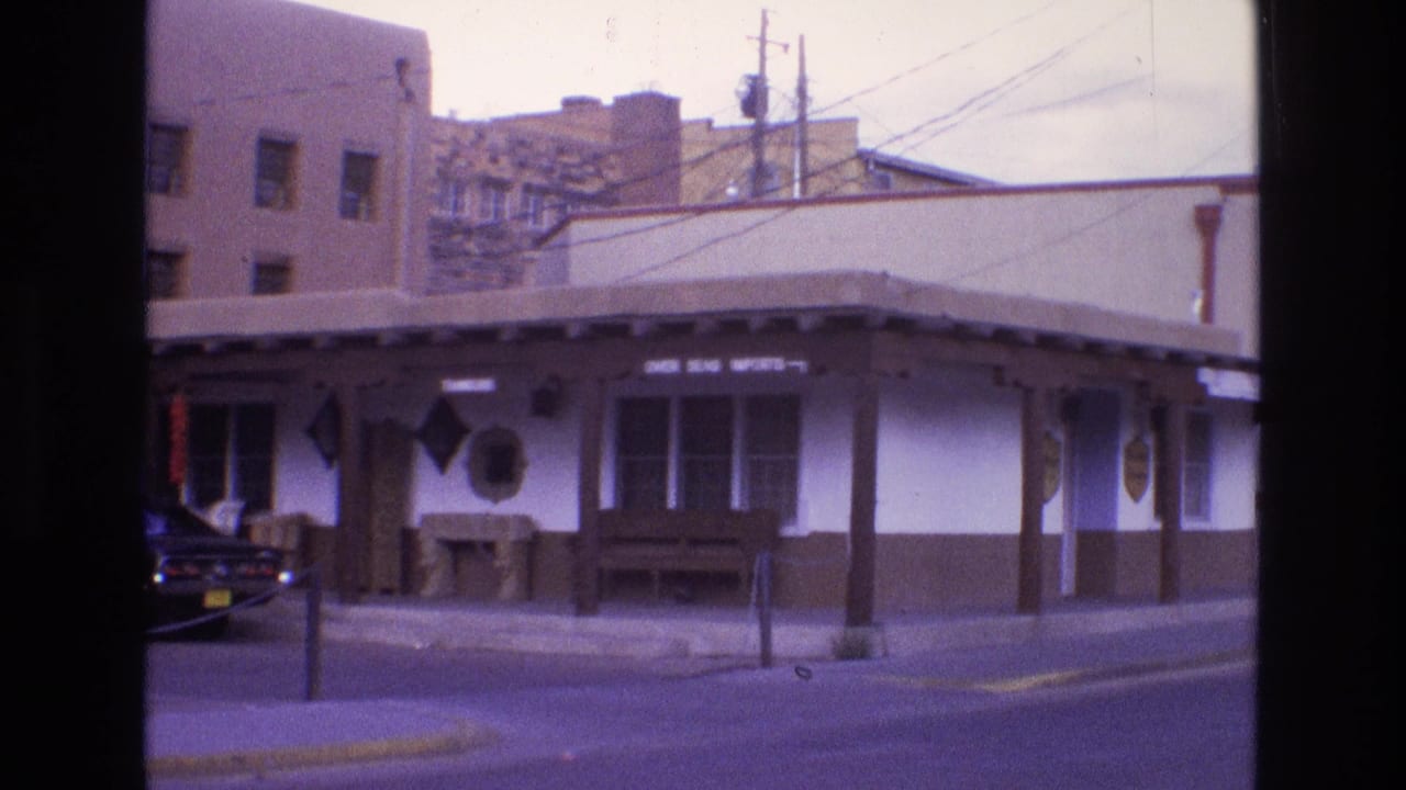 1973 New Mexico 8mm frame showing a street corner storefront, sidewalk posts, overhead utility lines, and a dark automobile partly visible at the curb.