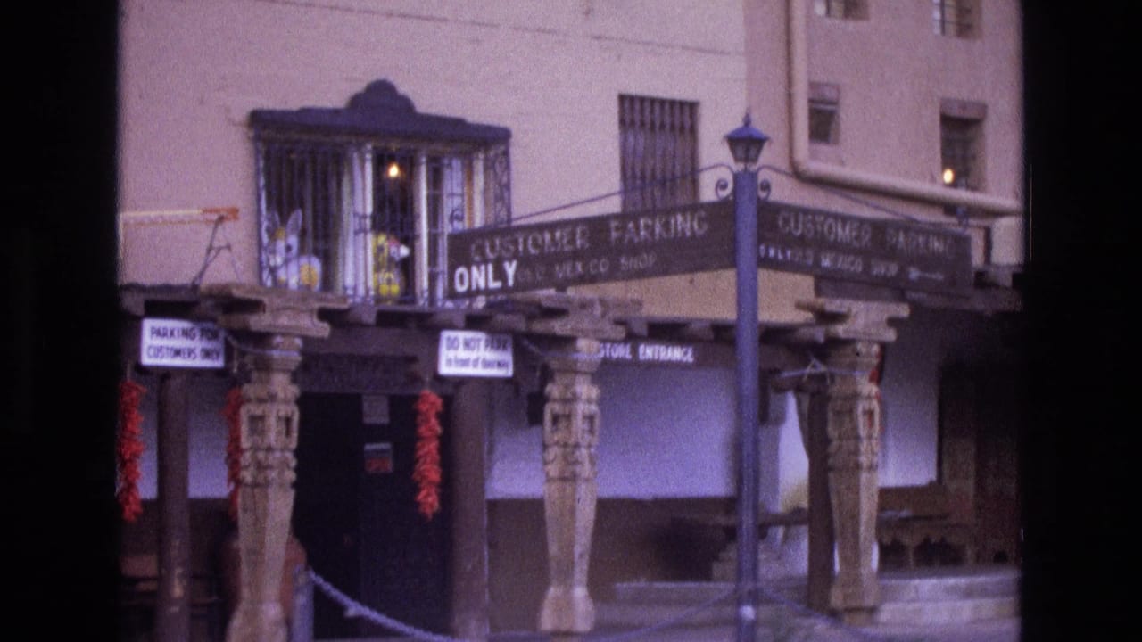 1973 New Mexico 8mm frame showing a covered storefront entrance with carved wooden posts, customer parking signage, barred windows, and a streetlamp in front.
