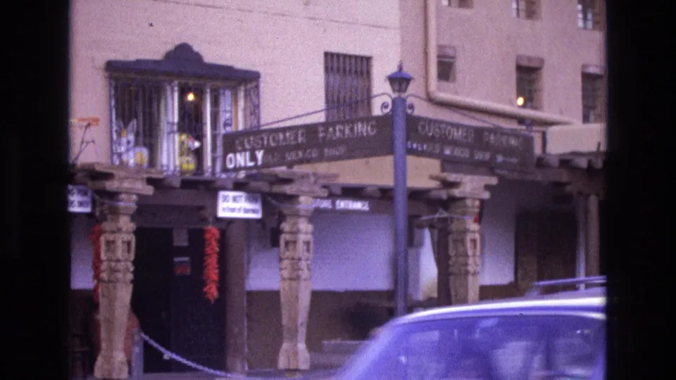 1973 New Mexico 8mm frame showing the decorated storefront entrance and signage while a light-colored car passes along the street in the foreground.