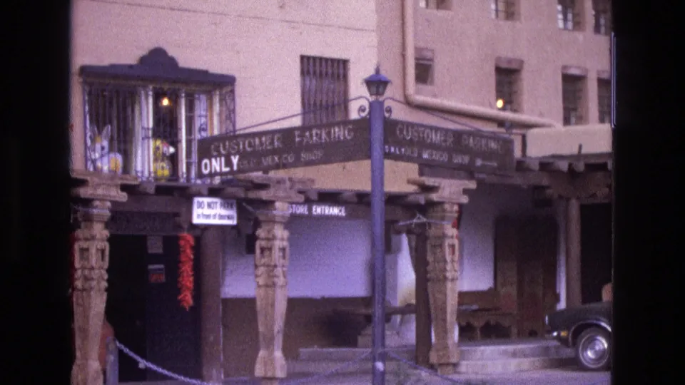 1973 New Mexico 8mm frame showing a Southwestern storefront with carved posts, customer parking signage, a streetlamp, and a dark car partly visible at the right edge.