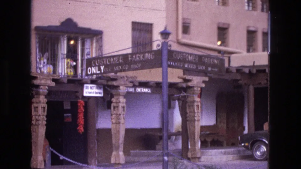 1973 New Mexico 8mm frame showing an Old Mexico Shop storefront with carved columns, customer parking signs, red chile strings, and a dark vintage car partly visible at the curb.