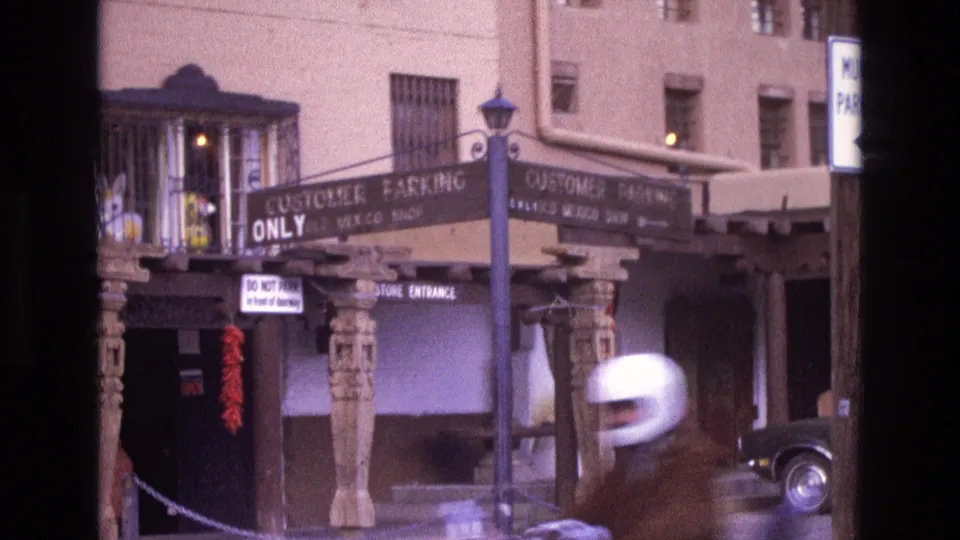 1973 New Mexico 8mm frame with a white-helmeted motorcyclist passing across the lower foreground in front of the Old Mexico Shop storefront, carved posts, and parking signs.