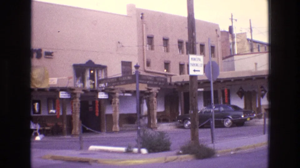 Wider 1973 New Mexico street view showing the Old Mexico Shop storefront with carved columns, customer parking signs, a dark parked car, sidewalk poles, and surrounding adobe-style building facades.