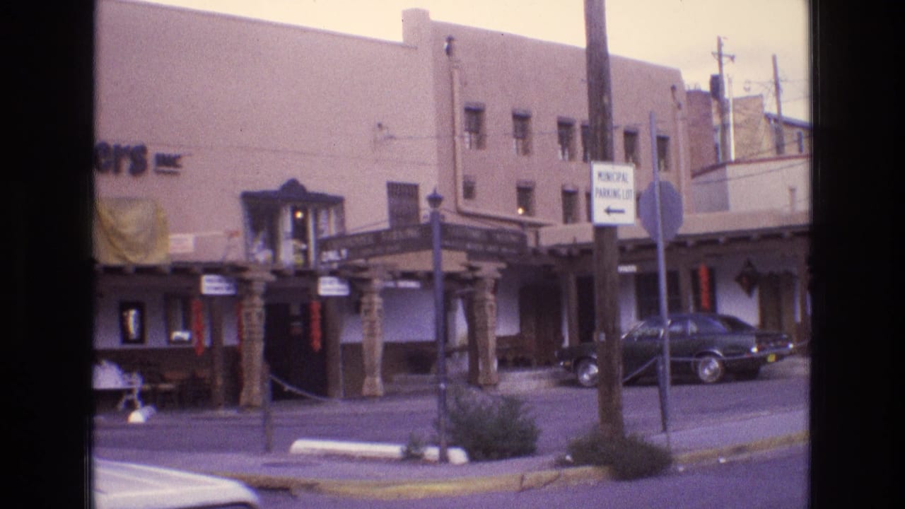 1973 New Mexico home-movie frame showing an adobe-style commercial building with a covered entrance, carved wooden posts, street signs, and a parked dark sedan.