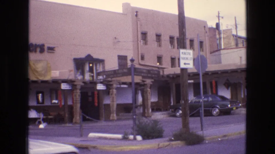 1973 New Mexico home-movie frame showing an adobe-style commercial building with a covered entrance, carved wooden posts, street signs, and a parked dark sedan.