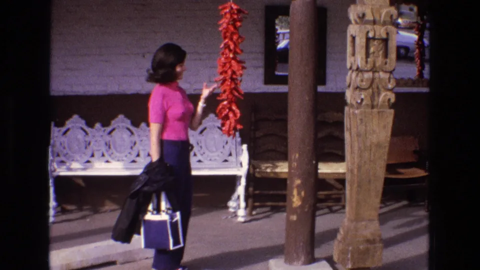 1973 New Mexico home-movie frame of a woman carrying a coat and bag while reaching toward a red chile ristra beside a white ornate bench and carved post.