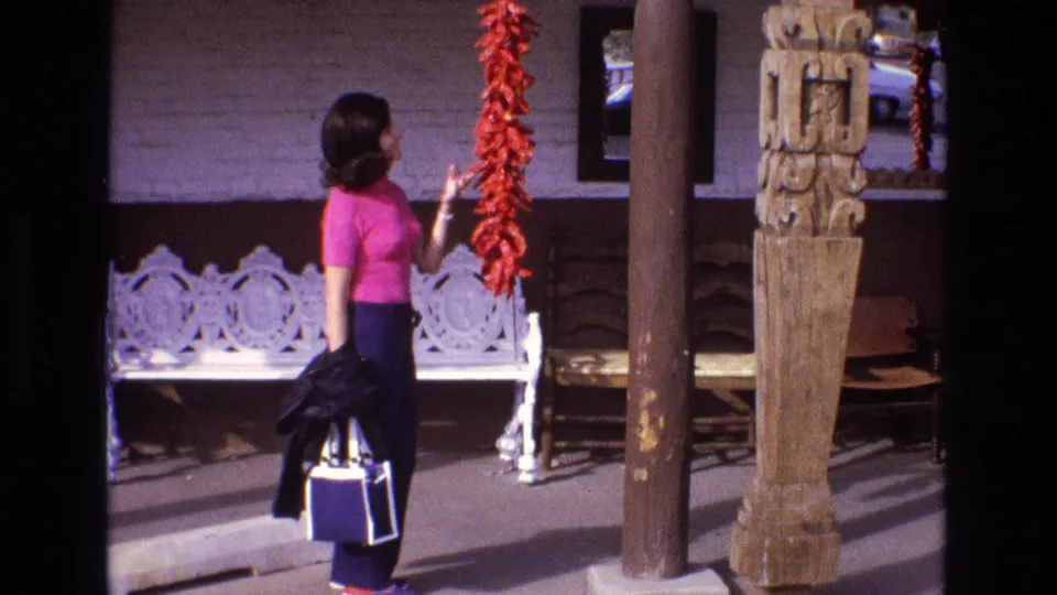 1973 New Mexico frame showing a woman beside a hanging red chile ristra, white bench, carved wooden columns, and a street-facing commercial exterior.