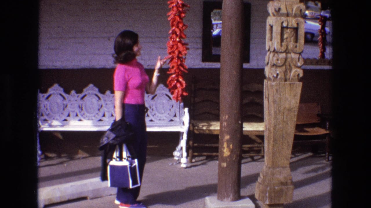 Woman in a pink shirt stands beside hanging red peppers on a porch.