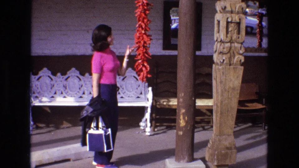 In 1973 New Mexico home-movie footage, a woman in a pink shirt and dark pants stands on a sunlit porch beside a long string of red chile peppers.