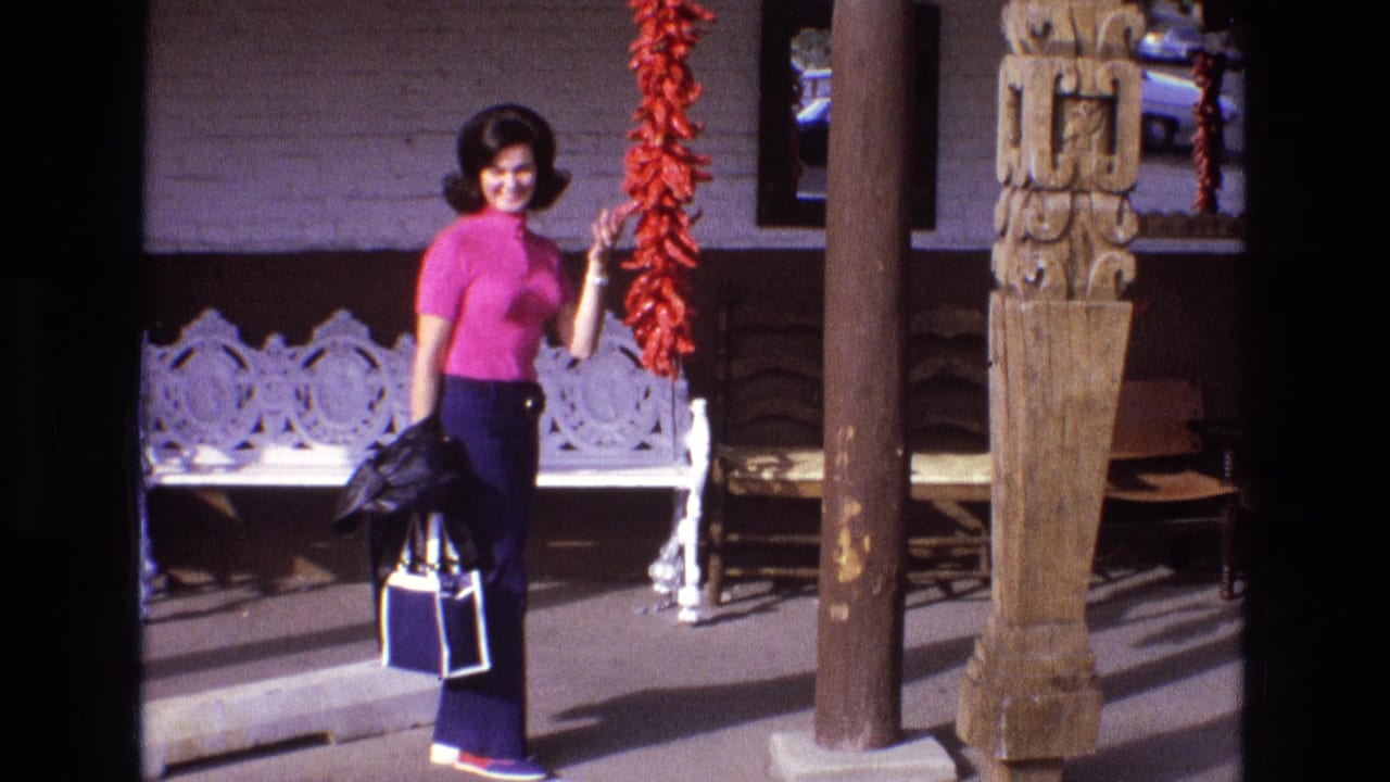 The woman smiles toward the camera, holding a jacket and blue-and-white bag while standing next to the hanging red peppers on the porch.