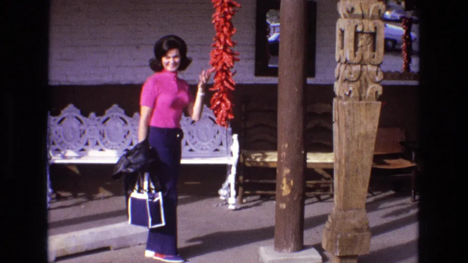 The woman smiles toward the camera, holding a jacket and blue-and-white bag while standing next to the hanging red peppers on the porch.