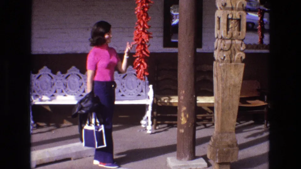 The woman reaches toward the red chile pepper string, with the white bench, porch post, and carved wooden feature visible behind her.