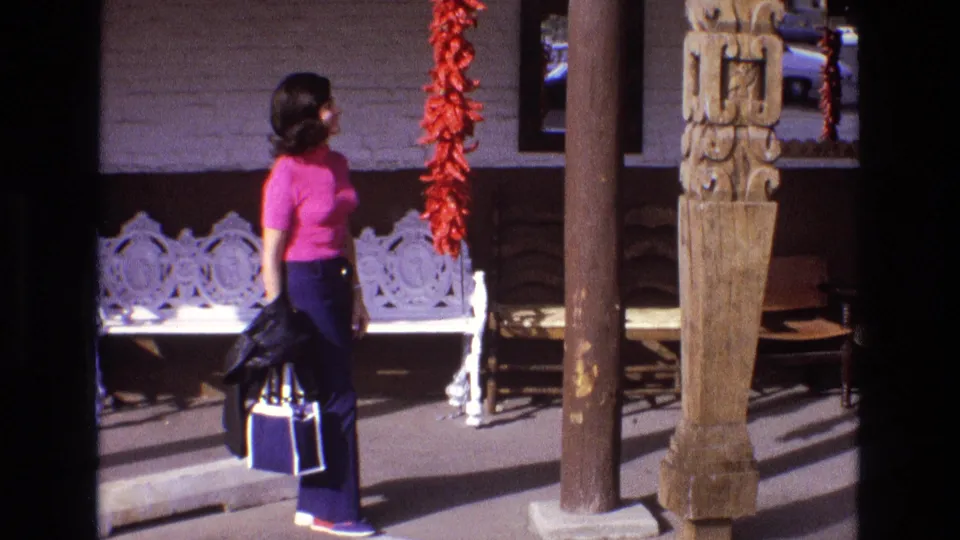 The woman turns her head toward the hanging red peppers, standing in bright sun with porch shadows, a carved post, and a white bench in view.