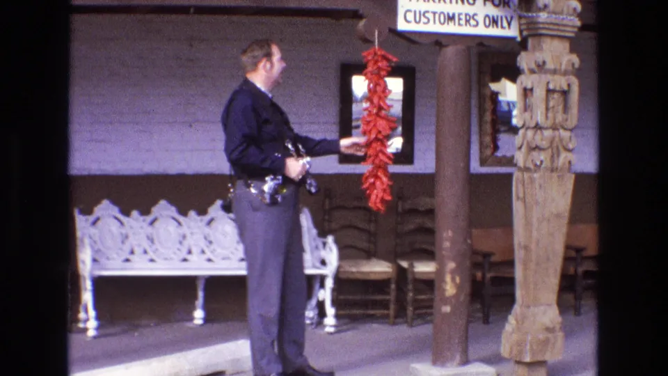 In supported 1973 New Mexico home-movie footage, a uniformed police officer stands outside a building and reaches toward a red chile ristra near wooden posts and a white bench.