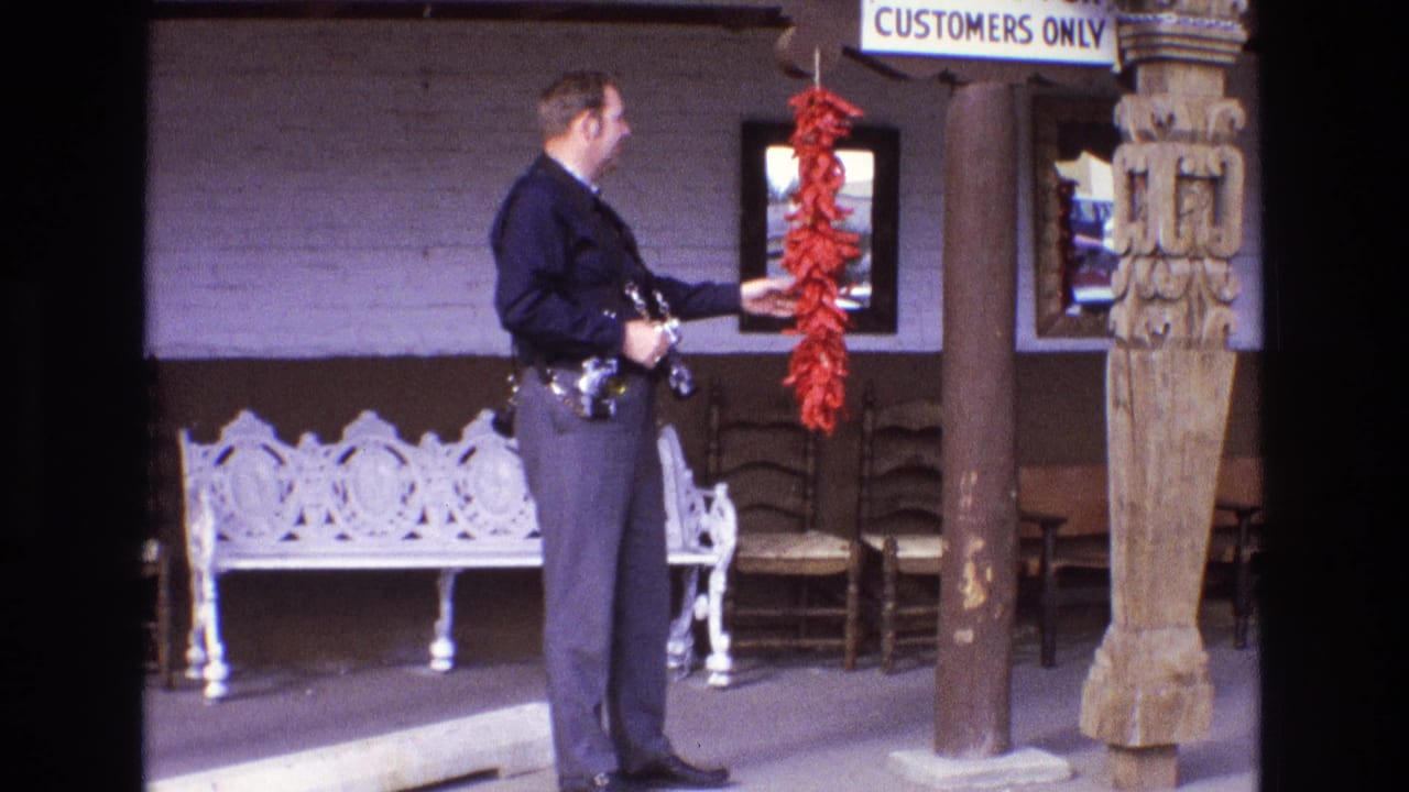 The uniformed officer stands in profile beside the hanging red chile peppers, with an ornate white bench, wooden chairs, and carved porch details visible behind him.