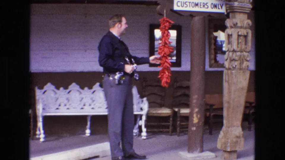 A 1970s uniformed police officer remains beside the red chile ristra, framed by a white metal bench, chairs, vertical wooden posts, and a partial customers-only sign.