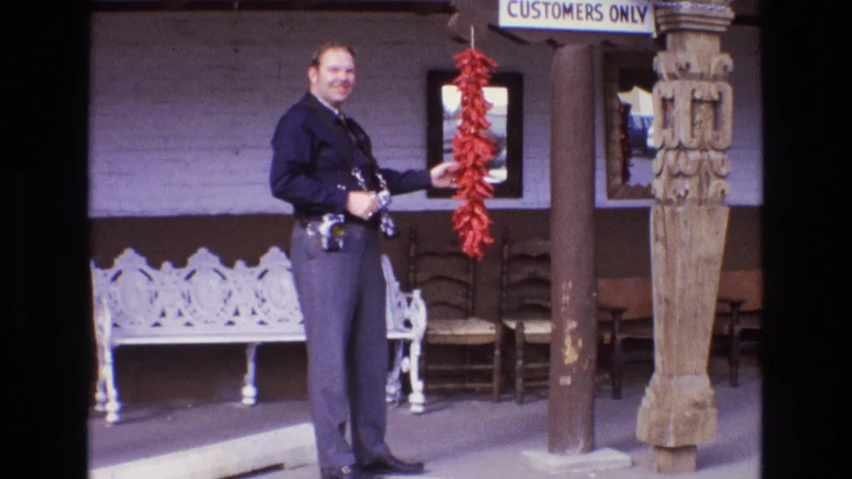The officer turns and smiles toward the home-movie camera while holding or touching the red chile string on a New Mexico building exterior.