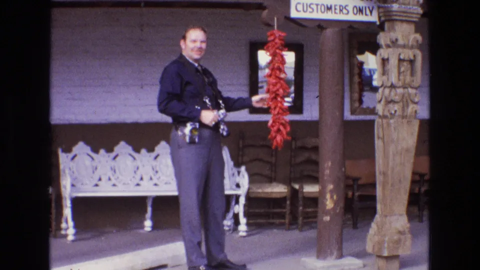 In 1973 New Mexico footage, a smiling uniformed man stands outside a business holding a string of red chili peppers near a white bench, wooden chairs, carved post, and customer parking sign.