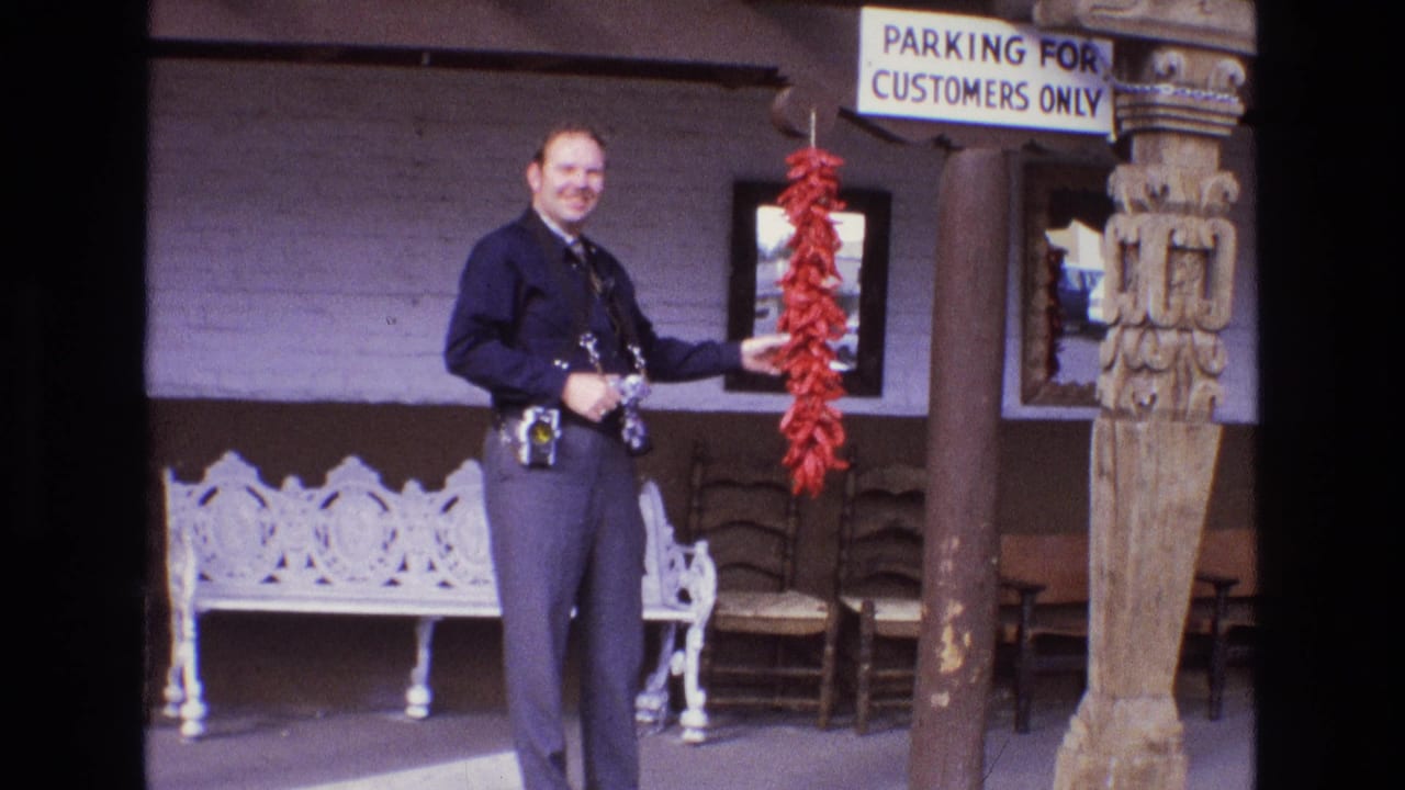 Smiling man in a dark uniform holding red chili peppers beside seating and a carved post.