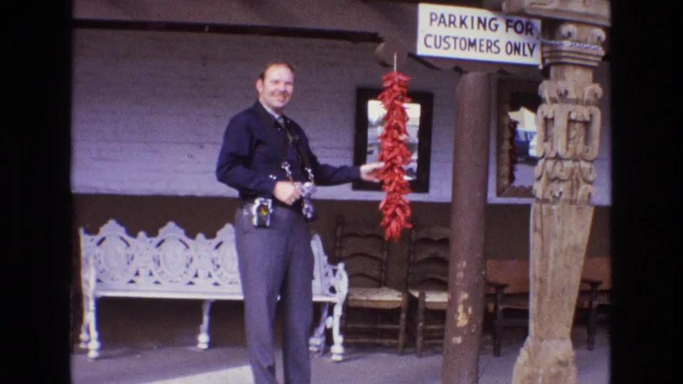 In the New Mexico home-movie frame, the man in a dark uniform smiles toward the camera while holding red chili peppers beside a carved post, bench seating, chairs, and a customer parking sign.