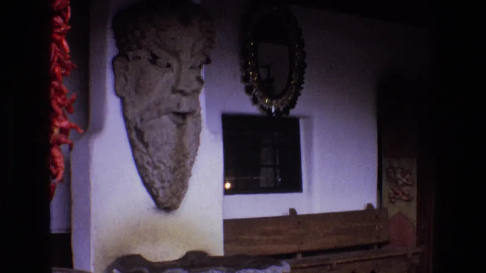 The frame shifts to a wall display with a large stone head sculpture, red chili peppers at the edge, a framed object or window, a wreath-like decoration, and a wooden bench below.