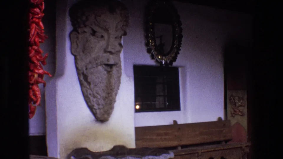 This New Mexico frame shows the stone head sculpture more clearly on a pale wall, with red chili peppers, a dark doorway, framed wall decor, and a long wooden bench in the foreground.