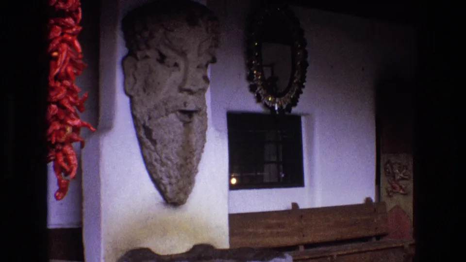 1973 New Mexico 8mm frame showing a weathered stone bearded head on a white plaster wall, with a red chile ristra at left, a round mirror, a small barred window, and wood below.