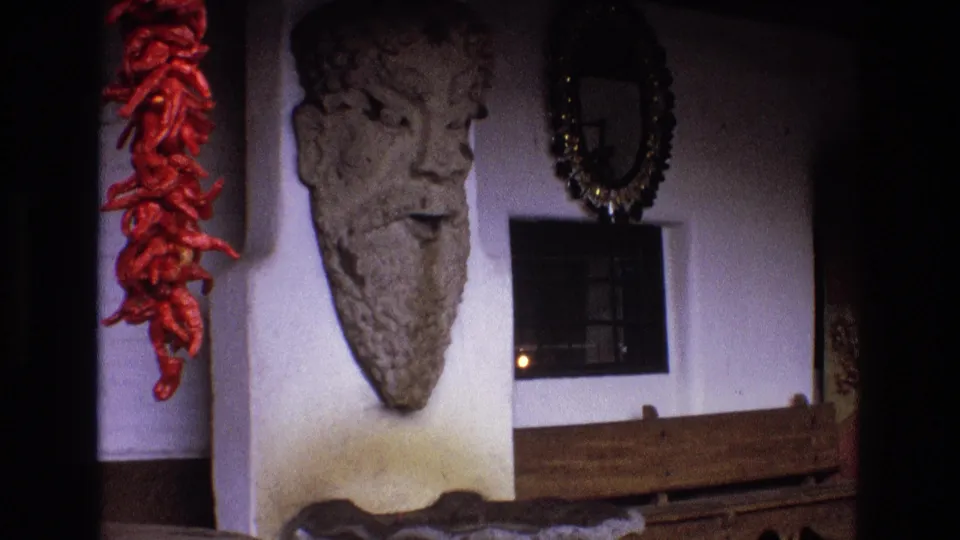 1973 New Mexico 8mm frame with the red dried chiles more prominent at left and a large rough-textured stone head mounted on the white wall near a mirror, window, and wooden element.