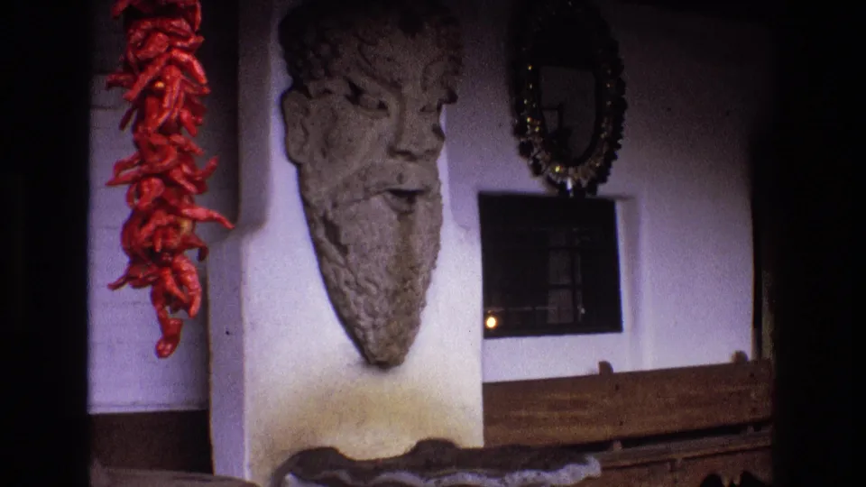 1973 New Mexico 8mm frame showing the final held view of the bearded stone wall sculpture, red chile ristra, circular mirror, small gridded window, and warm wood at the lower edge.