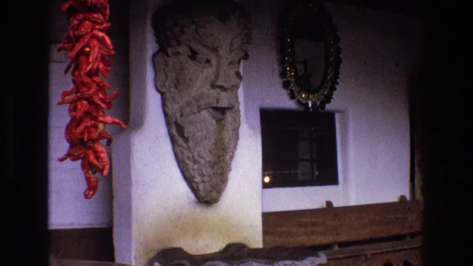 1973 New Mexico 8mm frame showing Southwestern trading post interior decor with a bearded stone face wall sculpture, hanging red chiles, an ornate mirror, and a wooden bench.