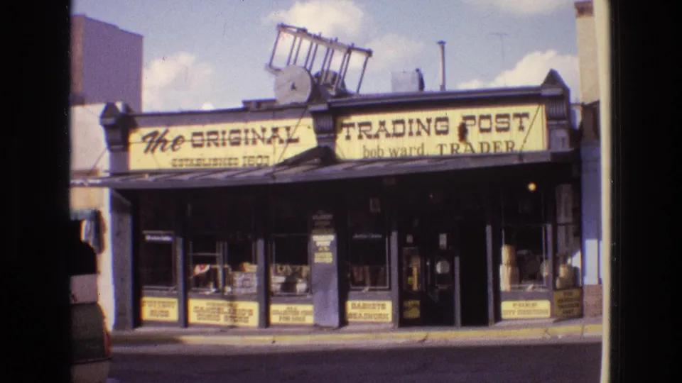 1973 New Mexico 8mm frame showing the trading post exterior with broad yellow signage, dark display windows, a doorway, and small shop signs along the facade.