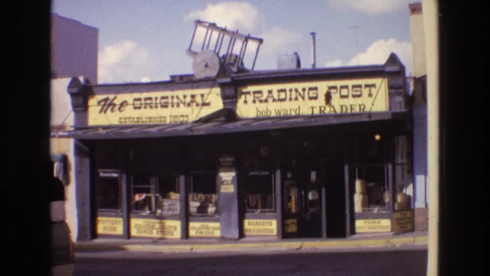 1973 New Mexico 8mm frame showing The Original Trading Post storefront with yellow signage, dark trim, sidewalk windows, and a ladder on the roofline.