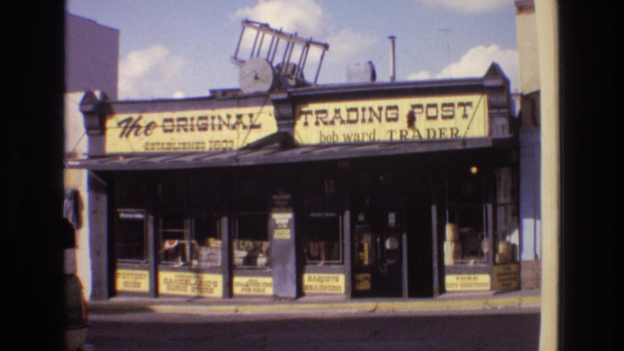 1973 New Mexico street view of a trading post storefront, with bold yellow lettering, glass display windows, and soft 8mm color and grain.