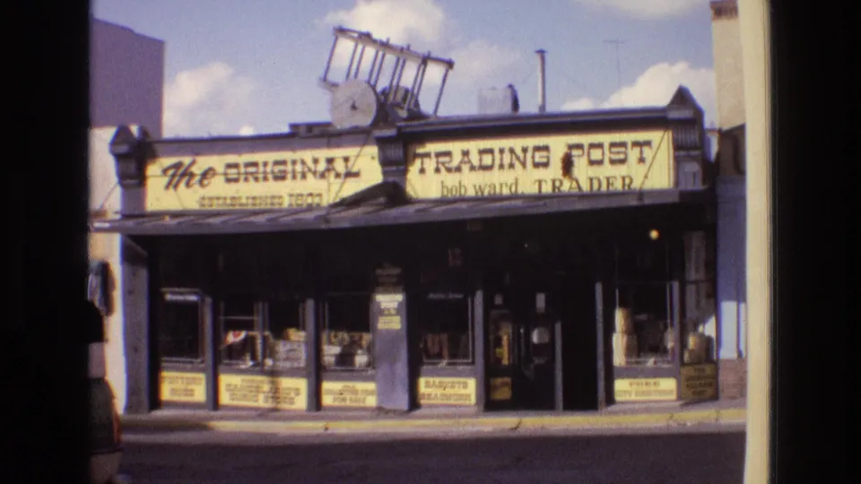 1973 New Mexico street view of a trading post storefront, with bold yellow lettering, glass display windows, and soft 8mm color and grain.