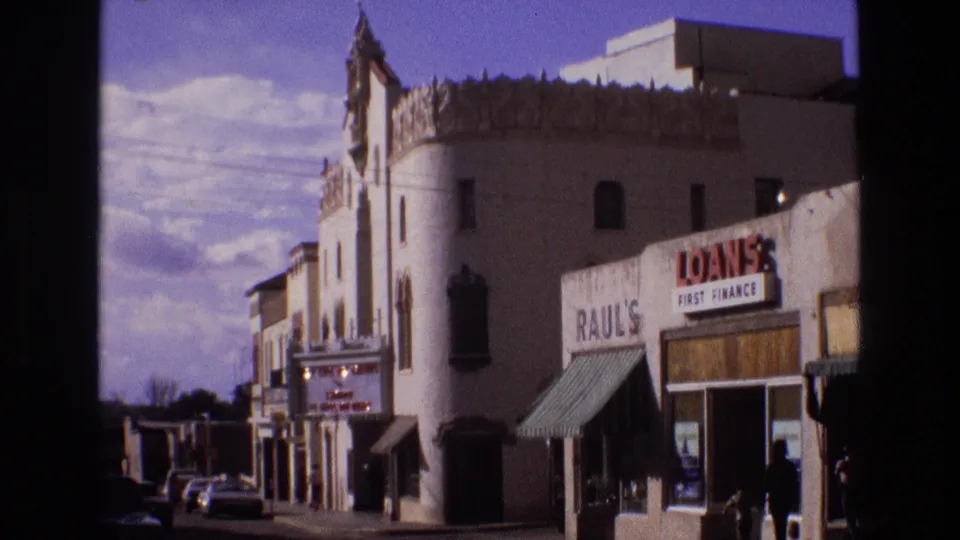 1973 New Mexico street frame showing a decorative theater-like building beside a loans storefront, with parked cars and small distant figures on the sidewalk.