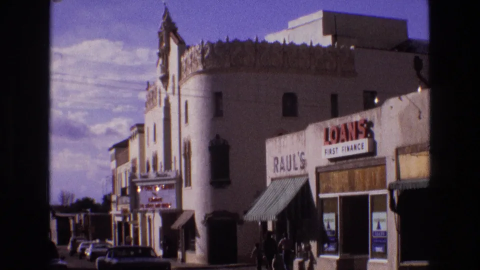 1973 New Mexico 8mm frame showing a white decorative building beside RAUL'S and LOANS FIRST FINANCE storefront signs, with parked cars along a sunlit small-town street.