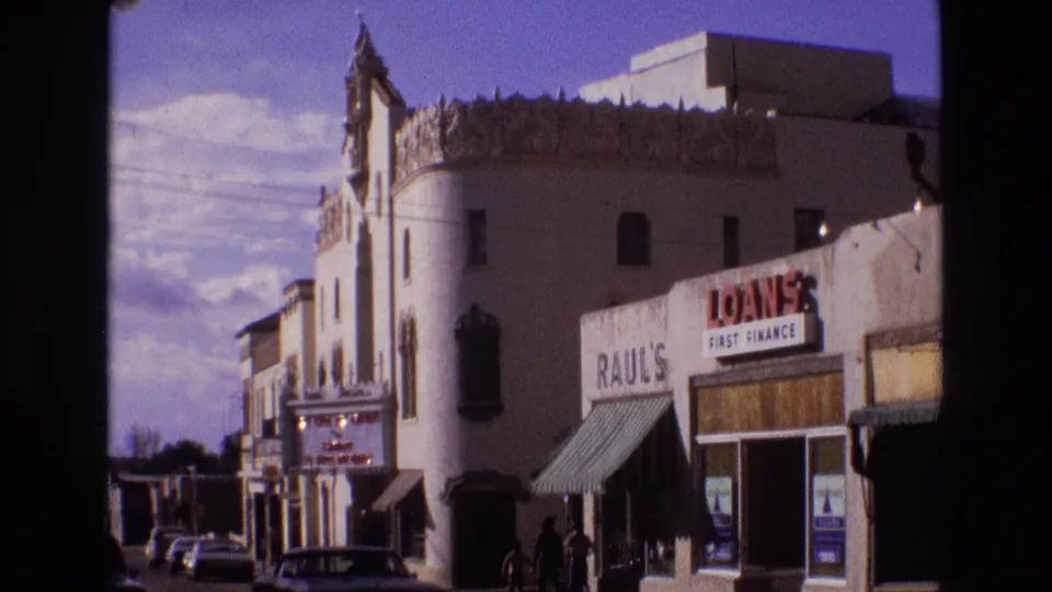 1973 New Mexico street frame with a white ornamental facade, visible business signage, parked cars, and a few people near the storefronts.
