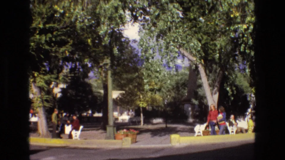 1973 New Mexico 8mm park view showing leafy trees, benches, planters, people in casual clothing, and small white animal figures or statues near the walkway.
