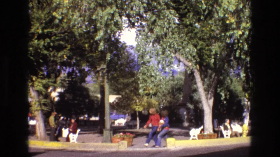 1973 New Mexico 8mm frame showing a tree-lined public park with seated visitors, white benches, planters, and people walking in casual clothing.