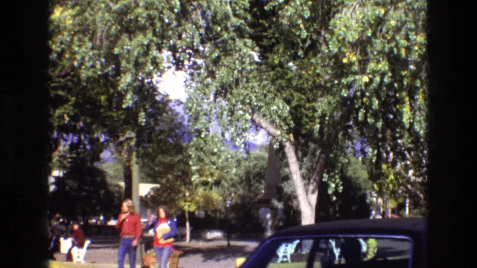 1973 New Mexico 8mm frame showing two women in casual clothes walking near benches and trees, with a dark car partly visible in the foreground.