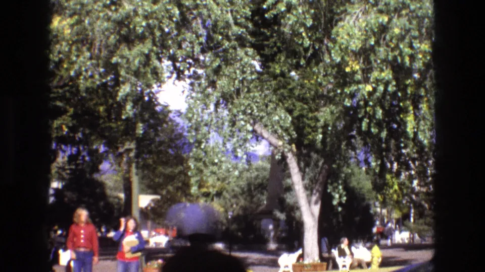 1973 New Mexico park frame with visitors seated on white benches, people walking in the middle distance, leafy trees overhead, and a blurred foreground passerby.