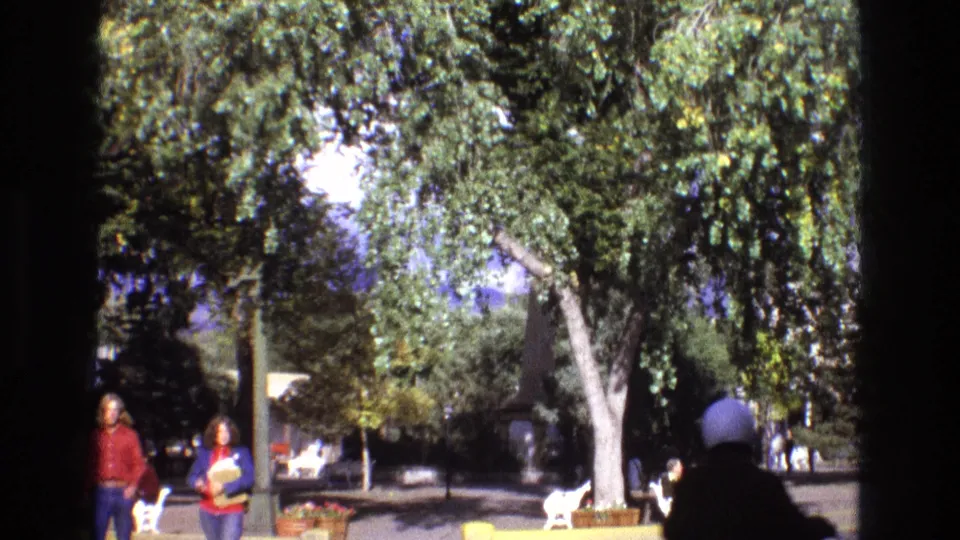 1973 New Mexico 8mm frame showing a shaded town square with leafy trees, distant people, benches, planters, and a white goat near the center of the plaza.