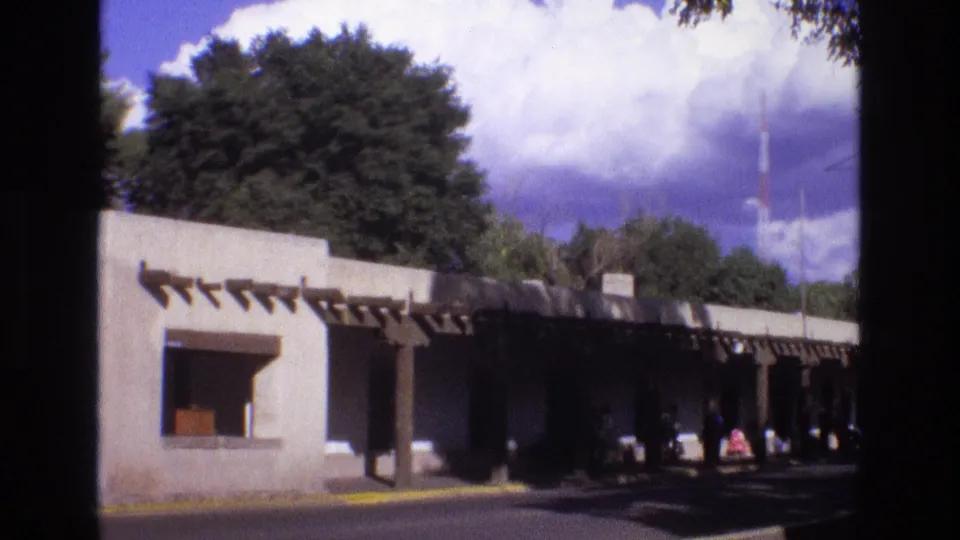 1973 New Mexico 8mm frame showing a low adobe-style building with wooden beam details, shaded walkway, trees, road edge, and cloudy blue sky.
