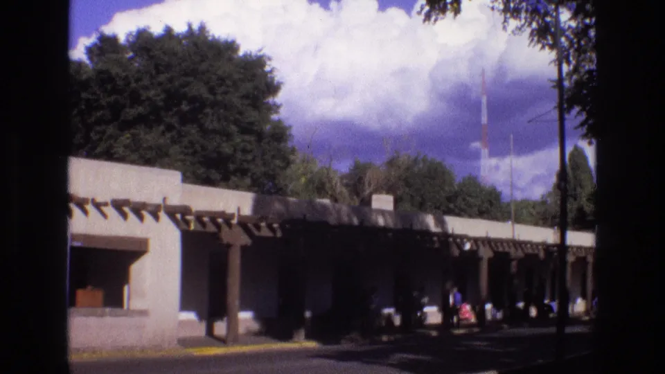 New Mexico 1973 home-movie frame of a low adobe-style structure with projecting beams, shaded pedestrian area, trees, and small distant figures near the walkway.