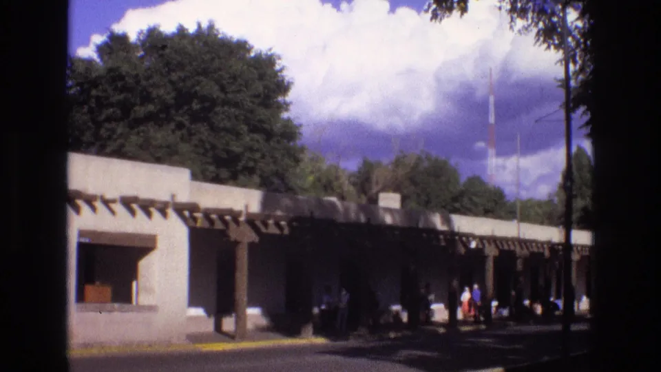 1973 New Mexico 8mm frame showing a low shaded building at the edge of a sunlit park, with trees, clouds, and small distant figures near the walkway.