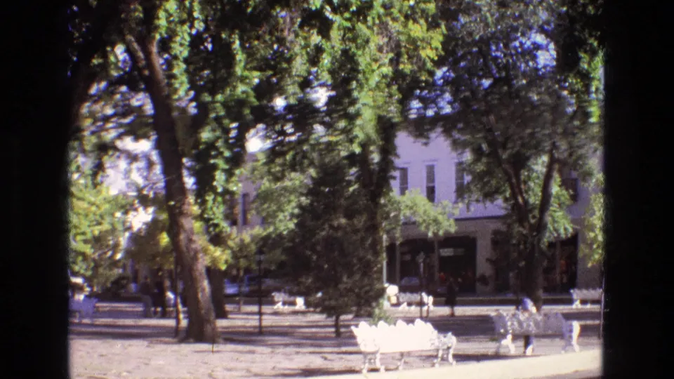 1973 New Mexico home-movie frame of white ornate benches under leafy trees, with a street, parked cars, and a two-story building in the background.