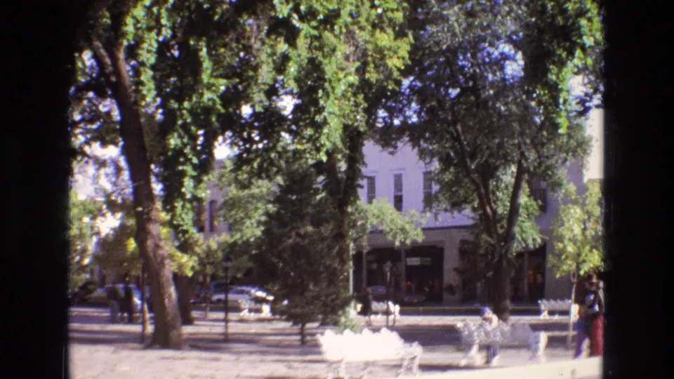 1973 New Mexico park scene with white benches, dense tree shade, a few distant pedestrians, and storefront-like building fronts across the street.
