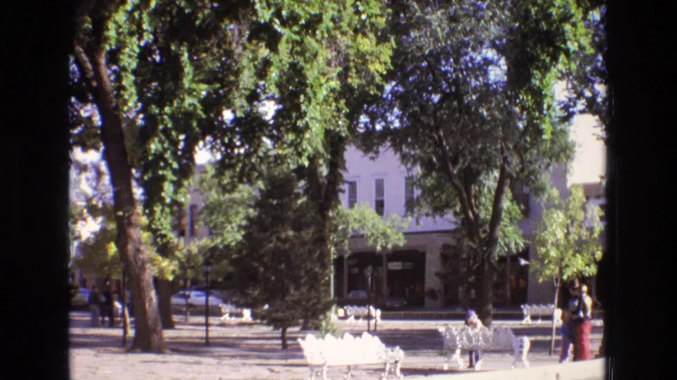 1973 New Mexico 8mm frame showing white ornate benches under leafy trees in a town park, with pedestrians and a street with parked cars in the background.