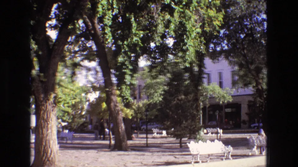 The final 1973 New Mexico frame shows white benches beneath dense trees, a person near the seating, and a quiet town street and buildings in the background.